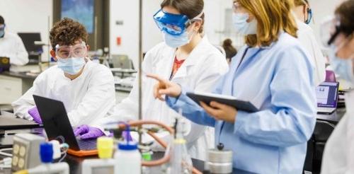 Four students wear lab coats, masks, and goggles while peforming a chemistry experiment. 